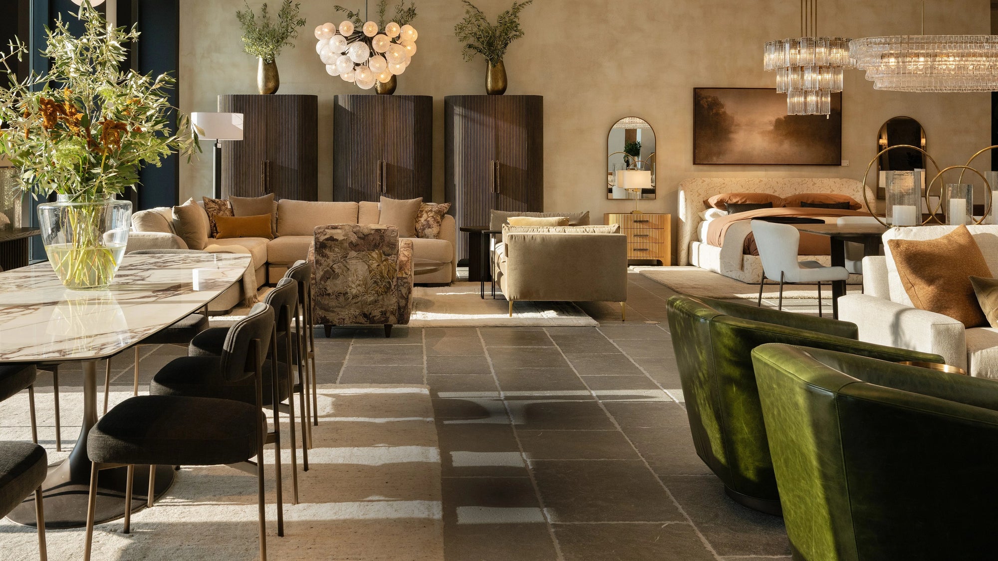 Arhaus showroom with stone dining table, neutral sectional, floral armchair, and green leather chairs in a sunlit, styled interior.