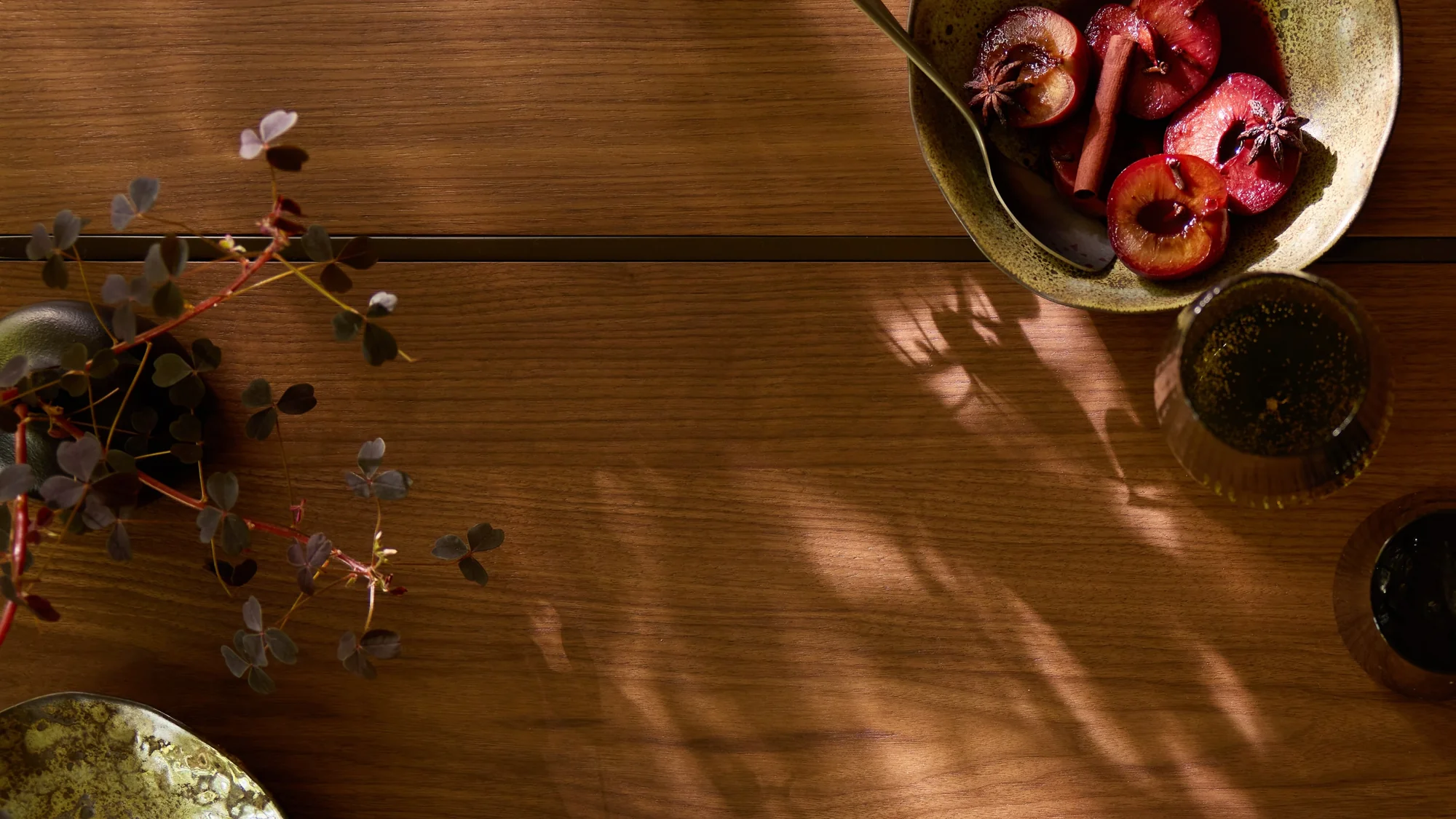 Overhead view of a wooden table with a bowl of halved plums spiced with star anise and cinnamon sticks, casting warm shadows in the light. A leafy plant with dark green clover-shaped leaves extends into the frame from the left, while two glasses, one with a dark drink and one with a light sparkling drink, sit near the bowl on the right.