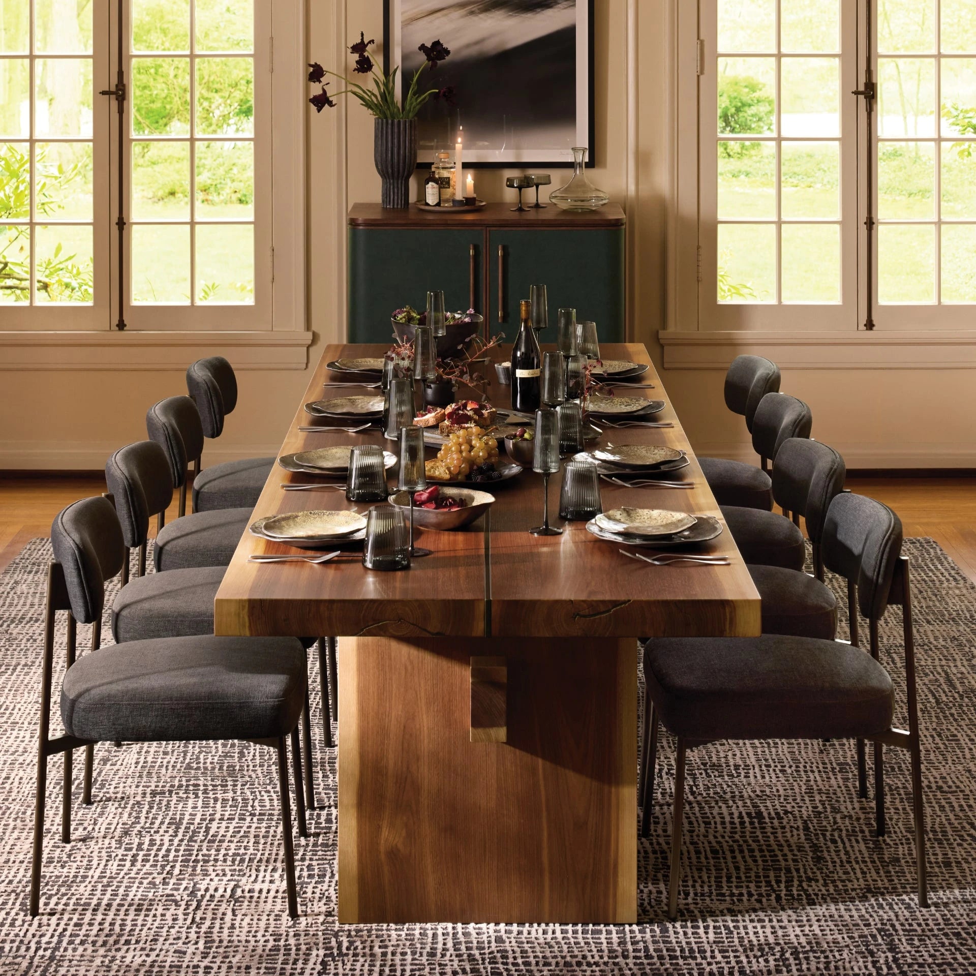 A long wooden dining table set for a dinner gathering with dark upholstered chairs, black glassware, and neutral-toned plates. The centerpiece features grapes, figs, and wine, creating an elegant, intimate setting framed by tall windows with garden views.
