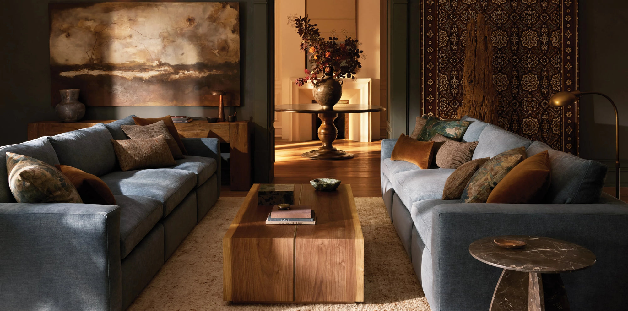 A warm, moody living room with two large blue sofas facing each other, accented by an array of textured throw pillows in earthy tones. A sleek wooden coffee table anchors the space, while abstract artwork, a patterned wall tapestry, and a round table with a dramatic floral arrangement add depth and elegance to the richly lit interior.