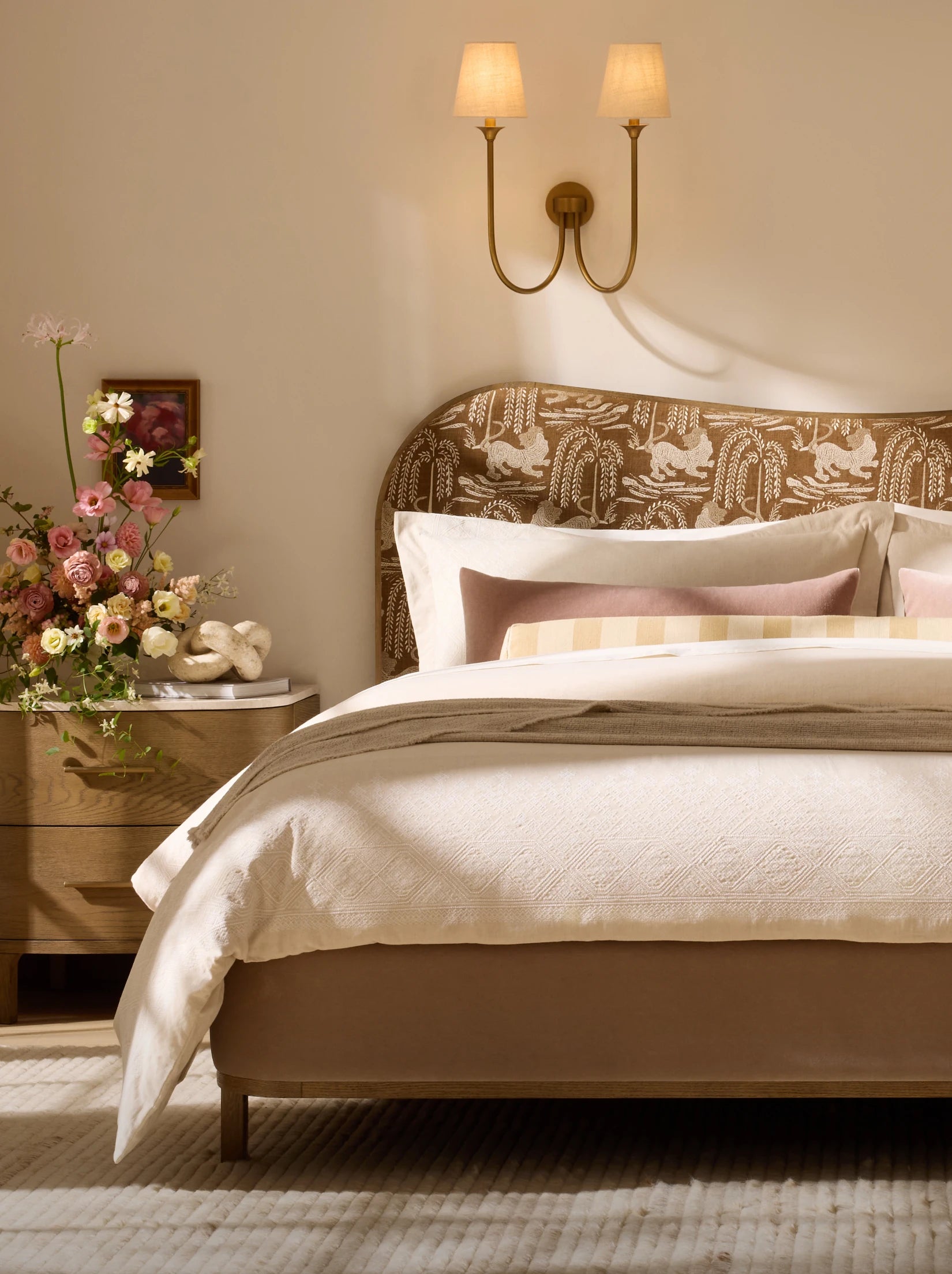 Bedroom with patterned headboard, layered neutral bedding, wall sconce, and nightstand with flowers.