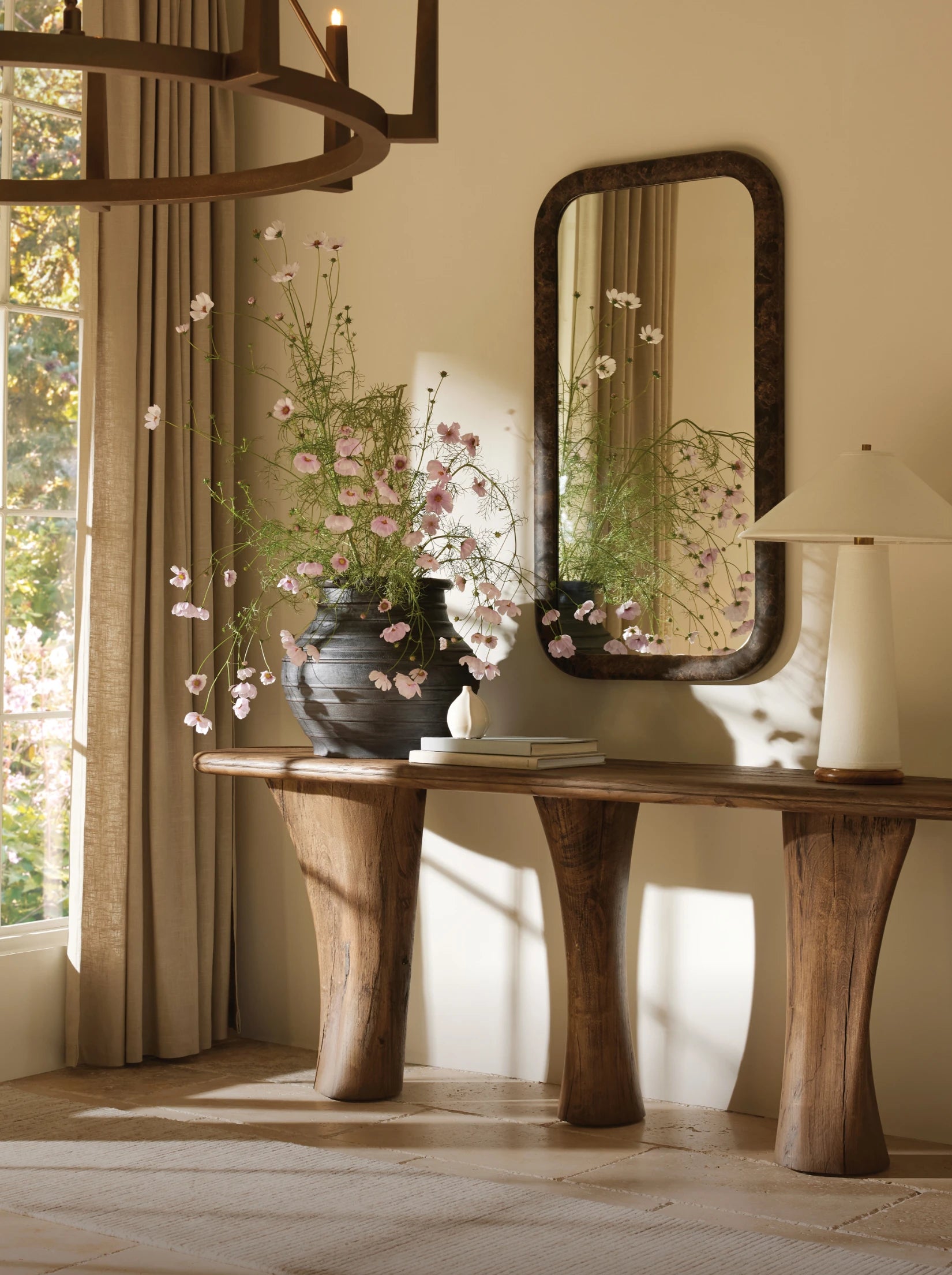 Entryway with wooden console table, large mirror, ceramic lamp, and vase of pink flowers.