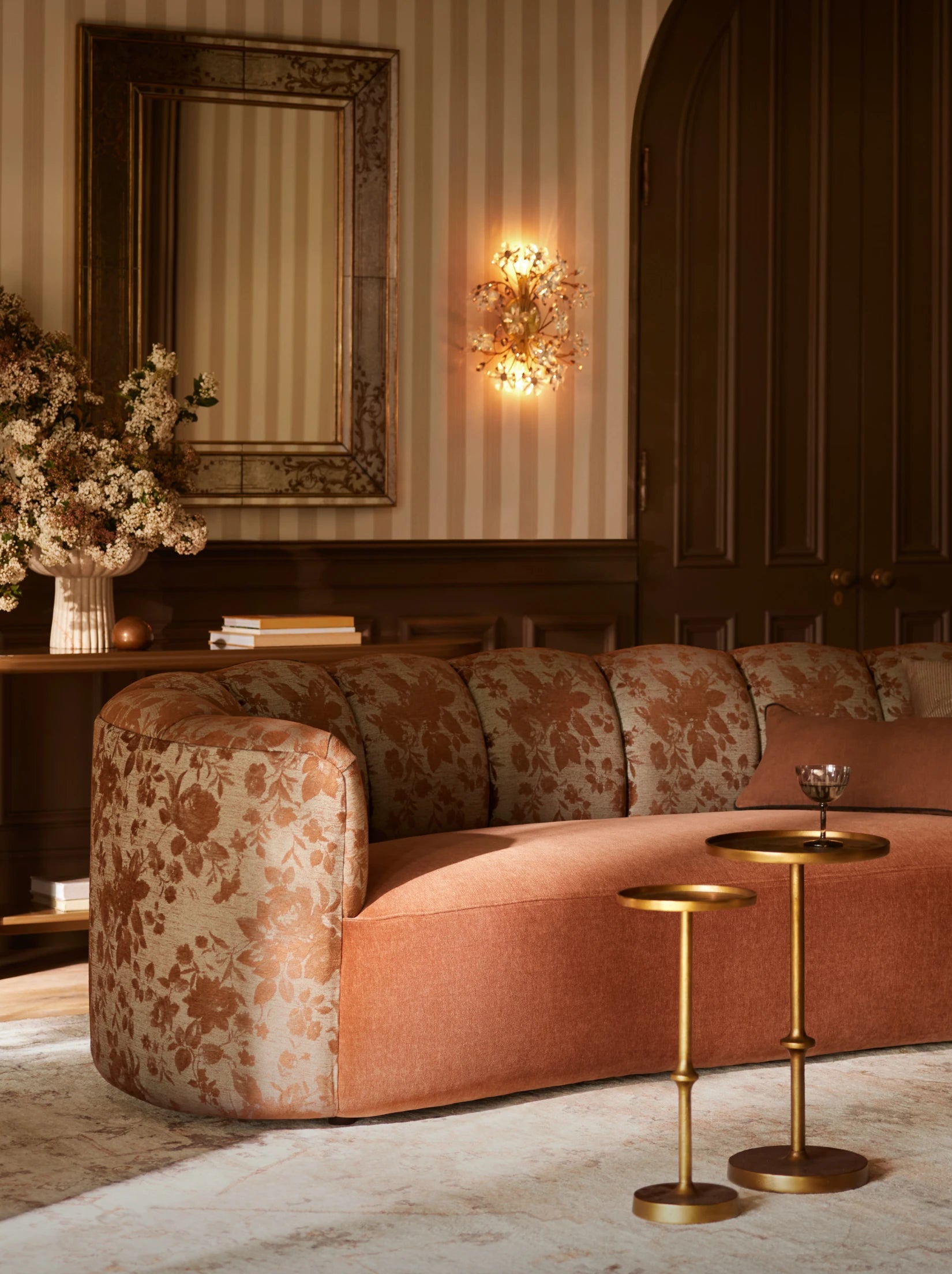 Room with floral-patterned curved sofa, gold side tables, wall mirror, and floral arrangement.