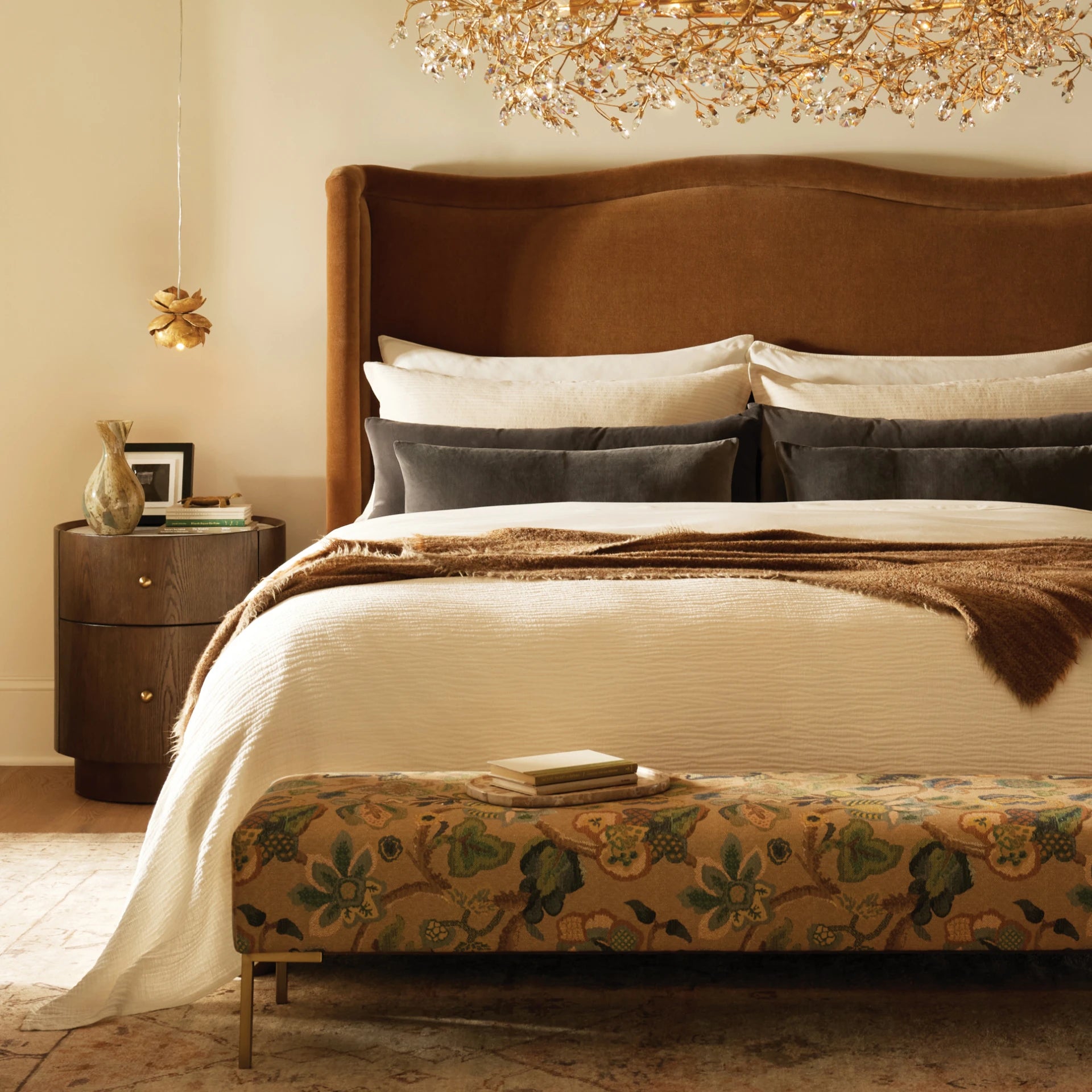 Bedroom with a tall brown upholstered headboard, layered neutral bedding, patterned bench, and gold chandelier.