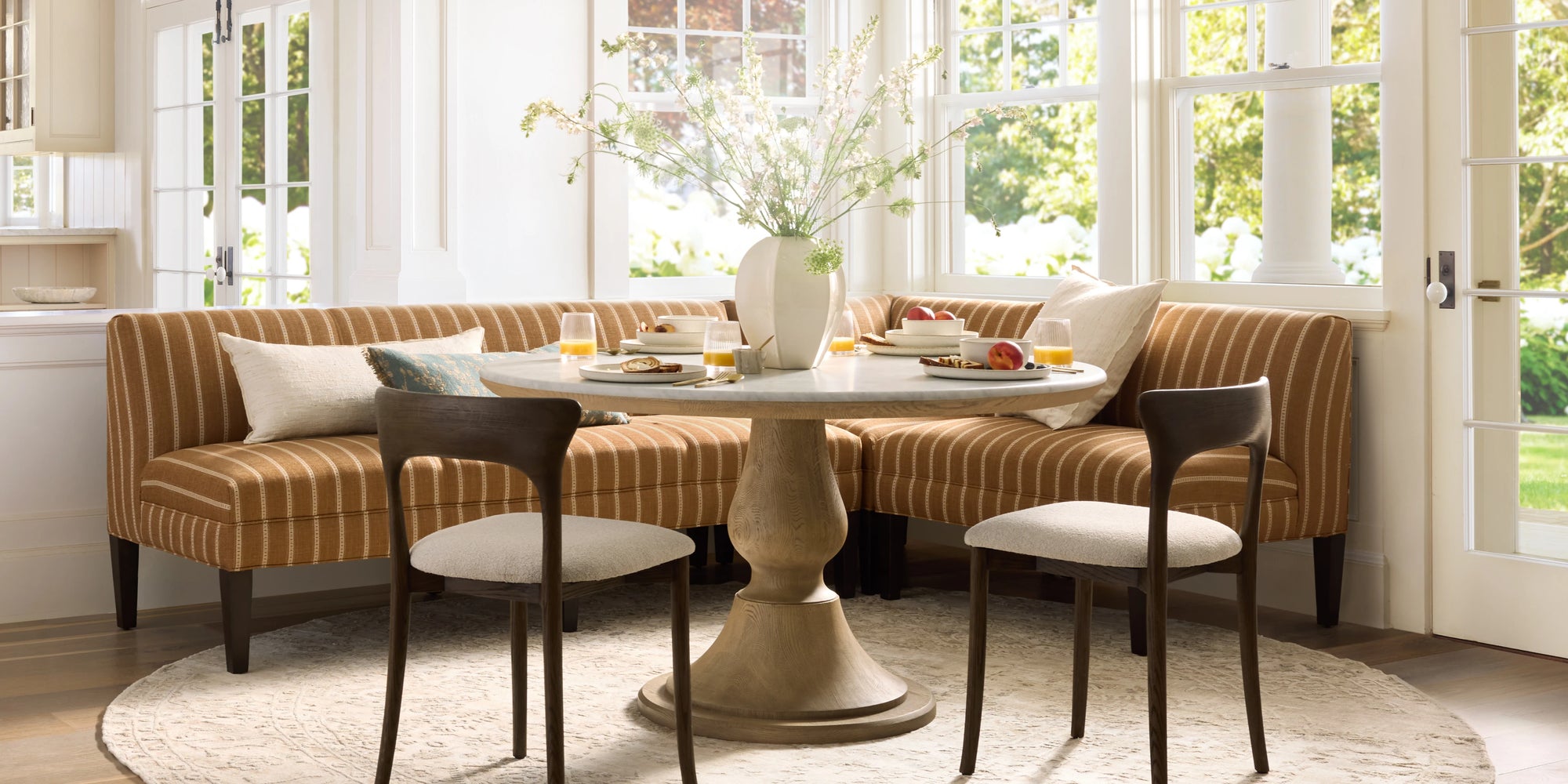 Sunlit dining nook with a striped banquette, round pedestal table, and wooden dining chairs beside large windows.