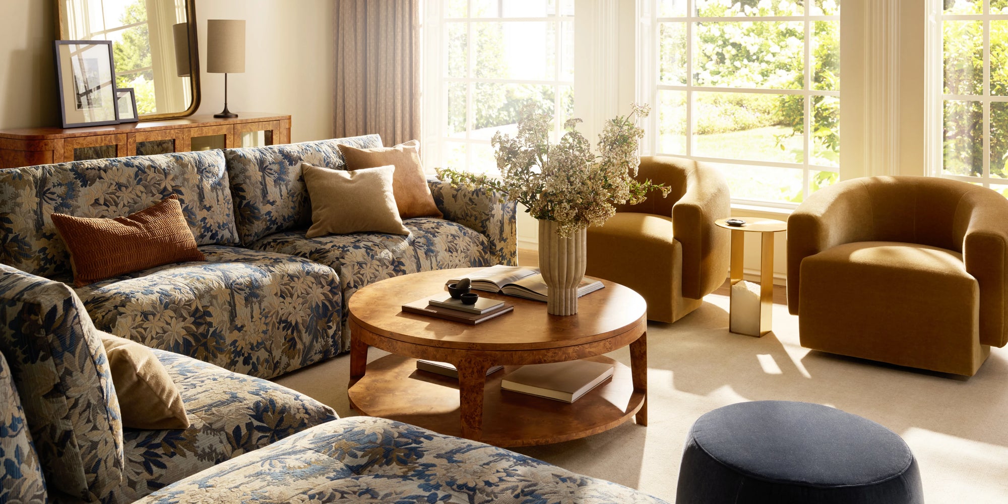 Sunlit living room with a floral-upholstered sectional, round wood coffee table, and upholstered accent chairs near large windows.