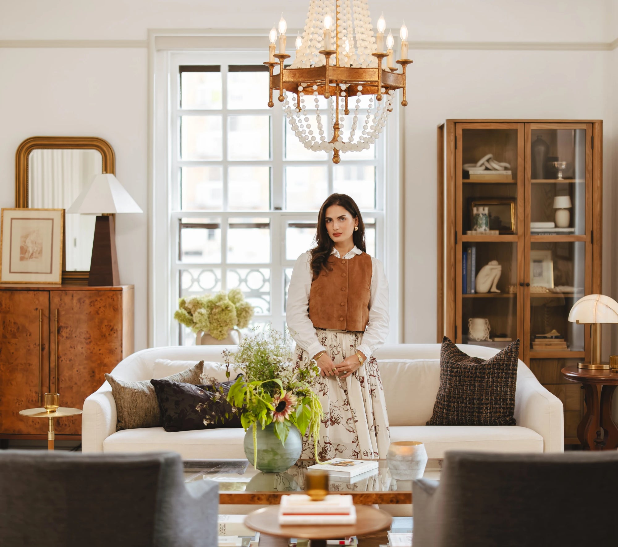 A styled living room with a modern classic design, featuring a white sofa layered with textured pillows, a wooden coffee table with fresh flowers, and a beaded chandelier overhead. A woman stands at the center, bringing warmth and personality to the bright, elegant space.