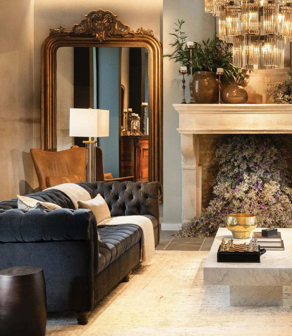 Luxurious Arhaus showroom featuring two navy tufted velvet sofas facing each other across a marble coffee table topped with decor books, a chess set, and a gold bowl. The space is styled with tall brass-framed mirrors, leather armchairs, modern floor lamps, and a grand crystal chandelier, all against a backdrop of neutral tones and lush greenery.