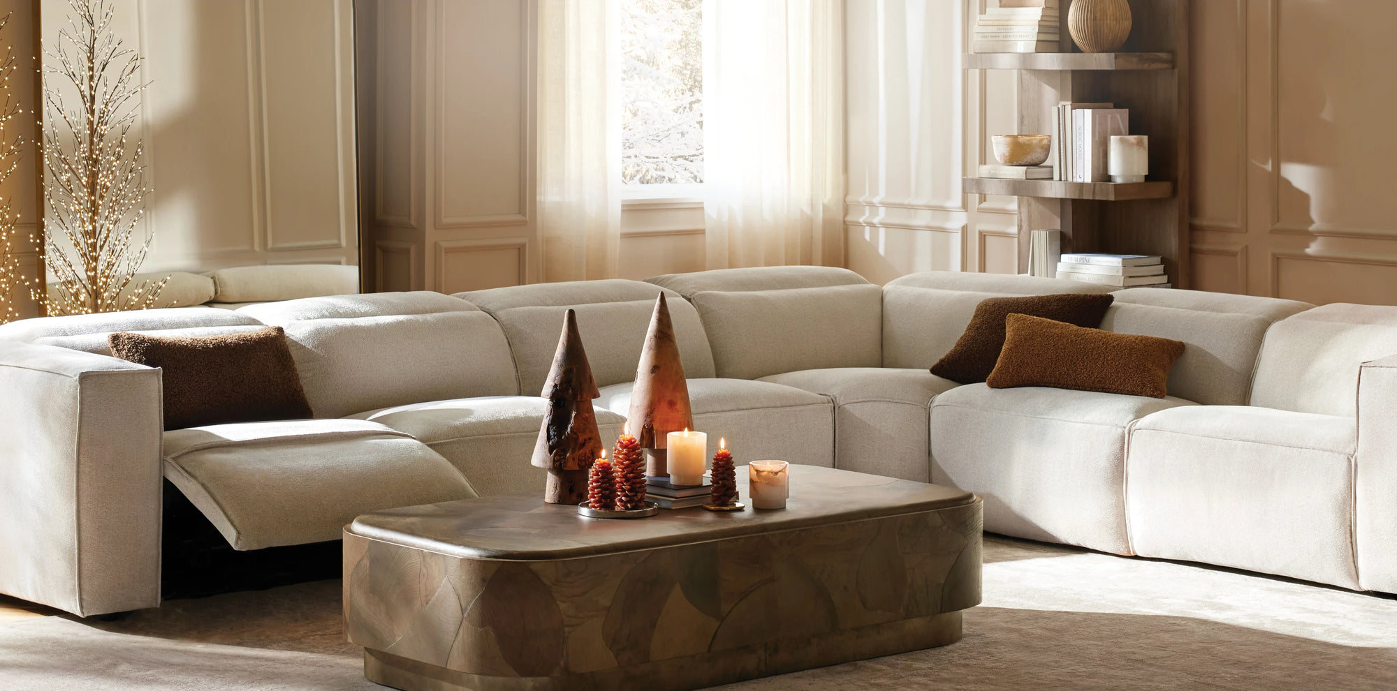 A spacious, cozy living room featuring a large cream-colored sectional sofa with built-in recliner, accented by brown textured pillows. A sculptural wood coffee table sits at the center, styled with wooden tree-shaped decor and glowing candles. Soft natural light filters through sheer curtains, highlighting wainscoted walls, a lit decorative branch, and a wooden shelving unit filled with books and ceramics.