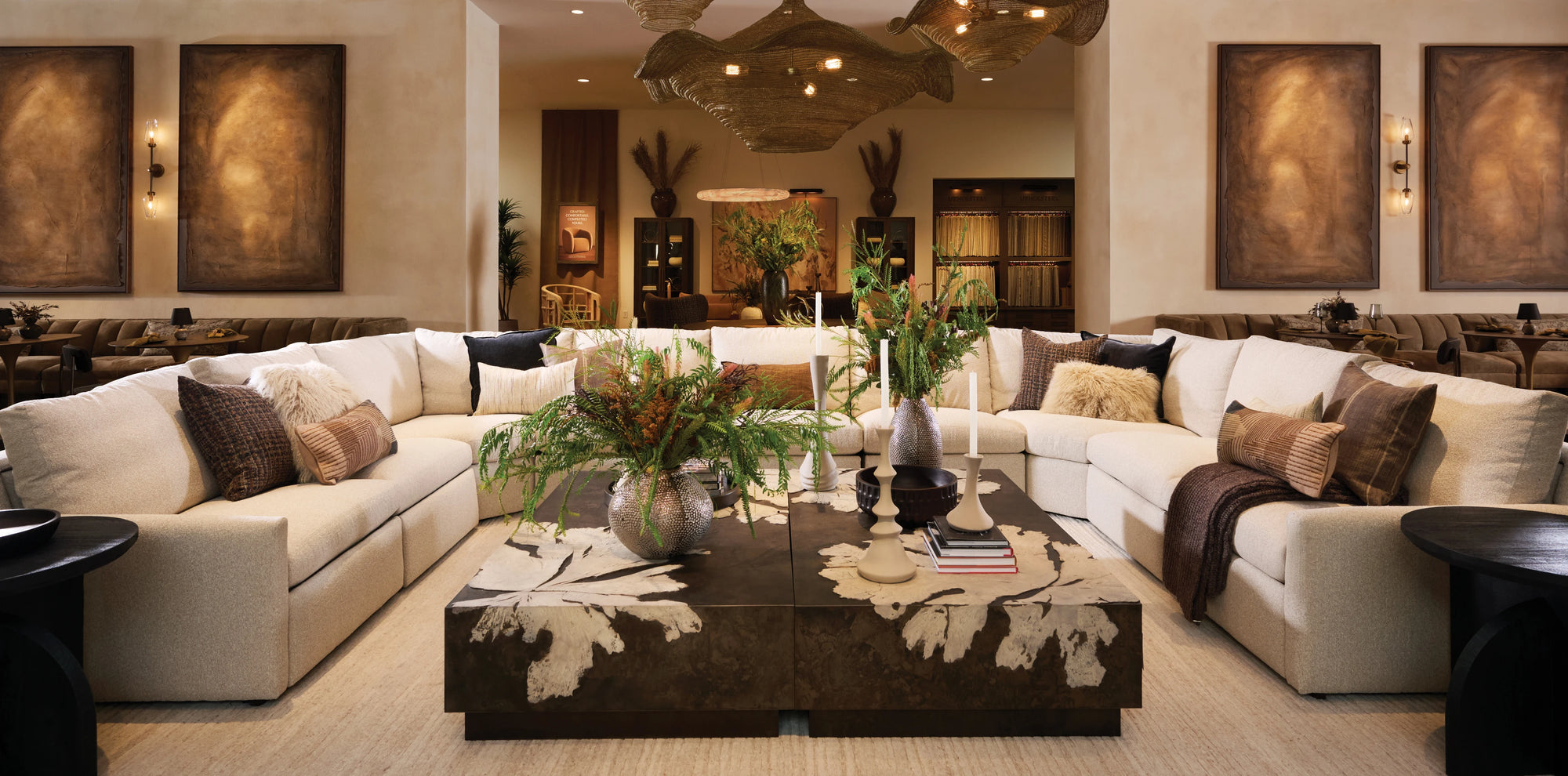 A spacious showroom living area featuring a large, cream-colored sectional sofa arranged in a U-shape, styled with textured neutral and earthy-toned pillows. At the center sits a pair of oversized square coffee tables with an abstract organic pattern, decorated with lush greenery in sculptural vases and tall modern candlesticks. Warm, ambient lighting, large wall art panels, and layered natural textures create an inviting, refined atmosphere.