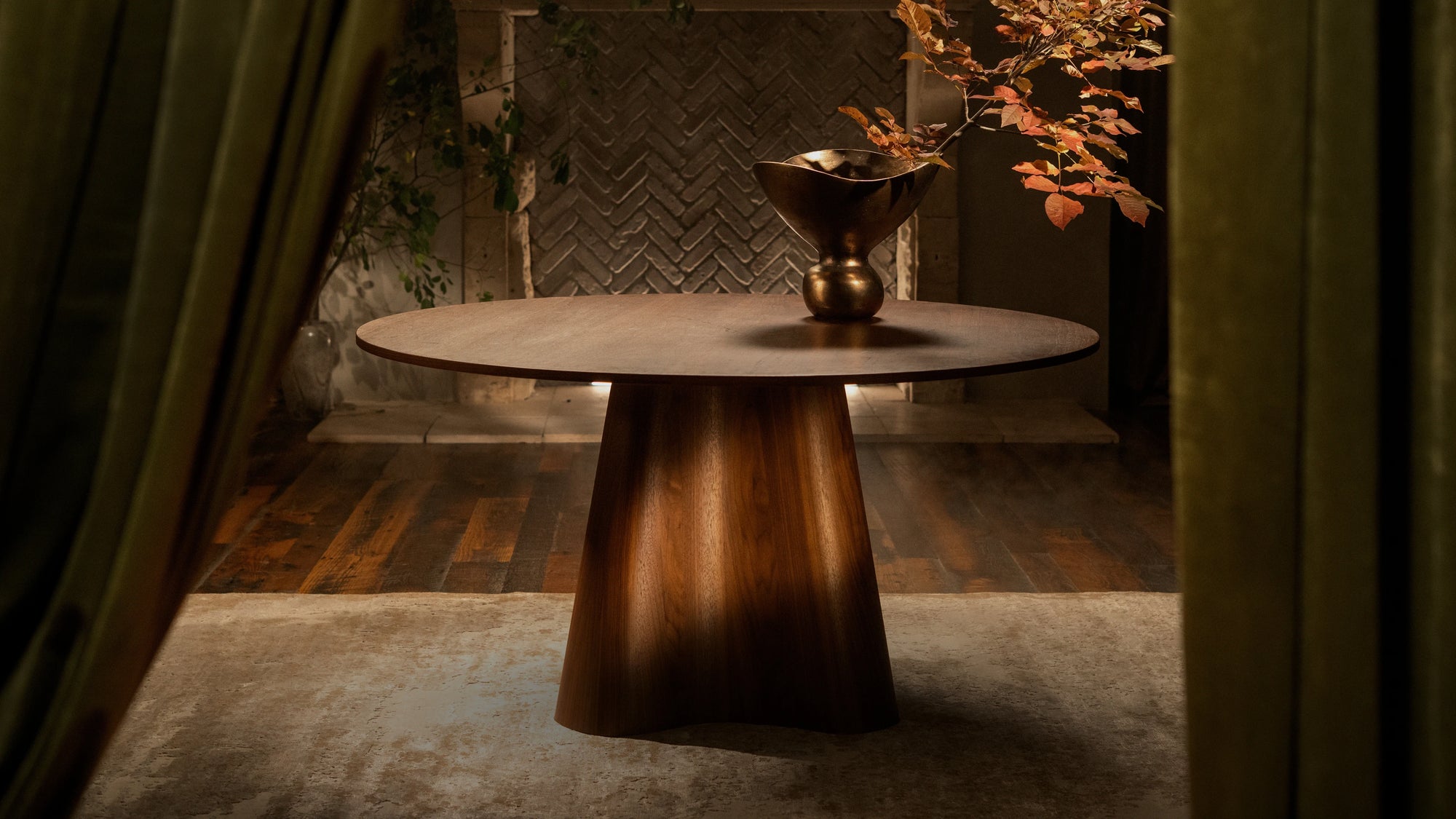 A modern round dining table with a sculptural wooden pedestal base, styled with brass decorative vessels and autumn foliage, set against a textured herringbone wall feature and flanked by rich olive green drapery in a sophisticated interior design setting.