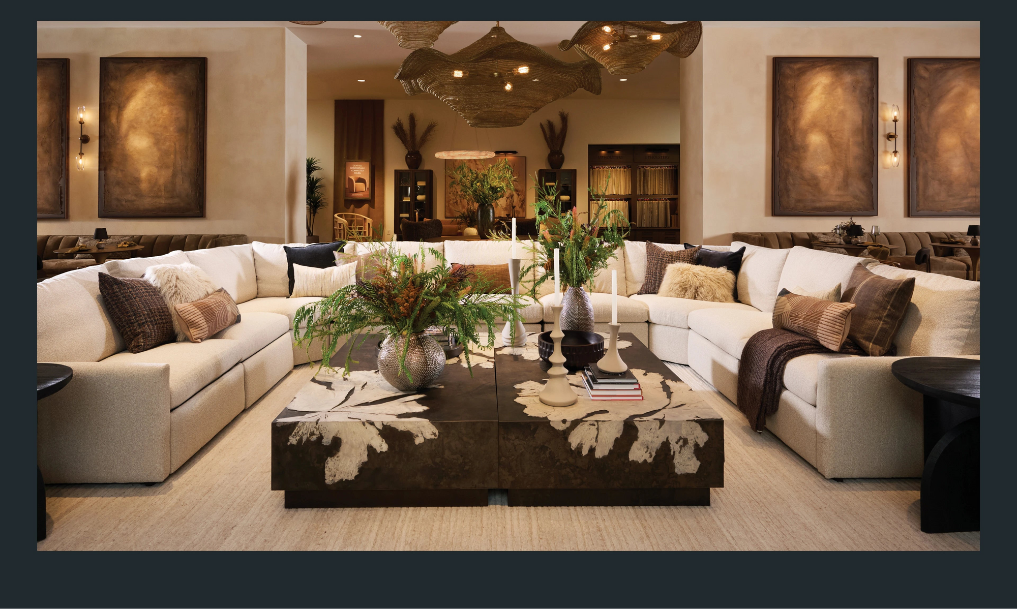 A spacious showroom living area featuring a large, cream-colored sectional sofa arranged in a U-shape, styled with textured neutral and earthy-toned pillows. At the center sits a pair of oversized square coffee tables with an abstract organic pattern, decorated with lush greenery in sculptural vases and tall modern candlesticks. Warm, ambient lighting, large wall art panels, and layered natural textures create an inviting, refined atmosphere.