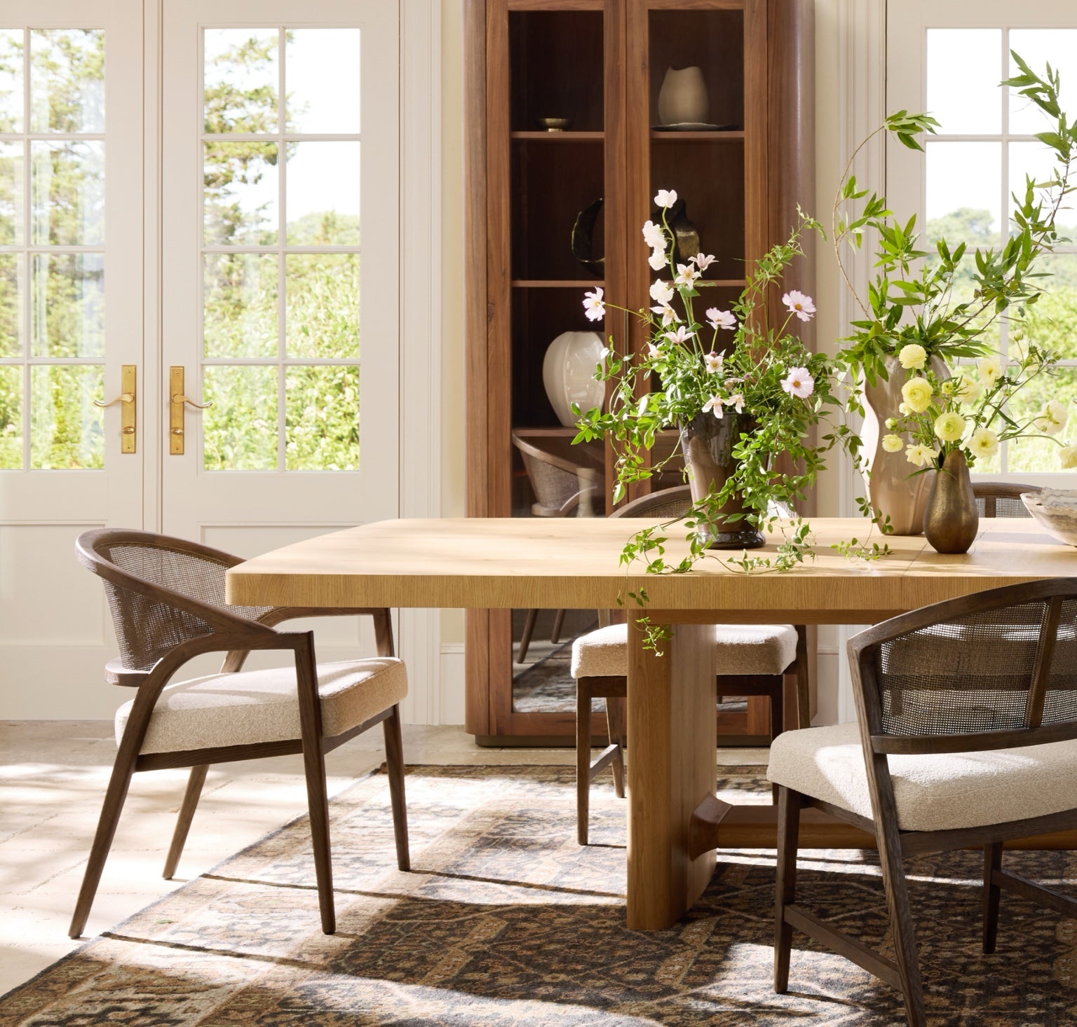 Sunlit dining room with a light wood rectangular table, upholstered chairs, and a tall wooden cabinet, styled with vases of fresh flowers and greenery near French doors overlooking lush outdoor foliage. 