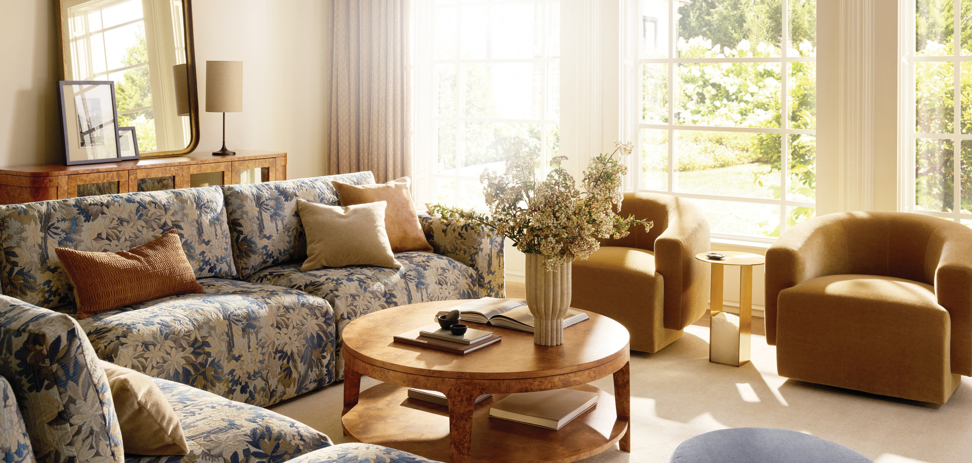 Bright, sunlit living room with a blue floral sofa, two warm-toned upholstered armchairs, and a round wooden coffee table styled with books and a vase of flowers, positioned beside large windows overlooking lush greenery.