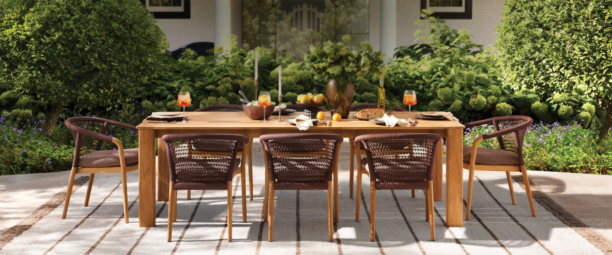 Outdoor dining space with woven dining chairs, a solid wood table, and seasonal tabletop styling, set among lush greenery in a sunlit garden setting.