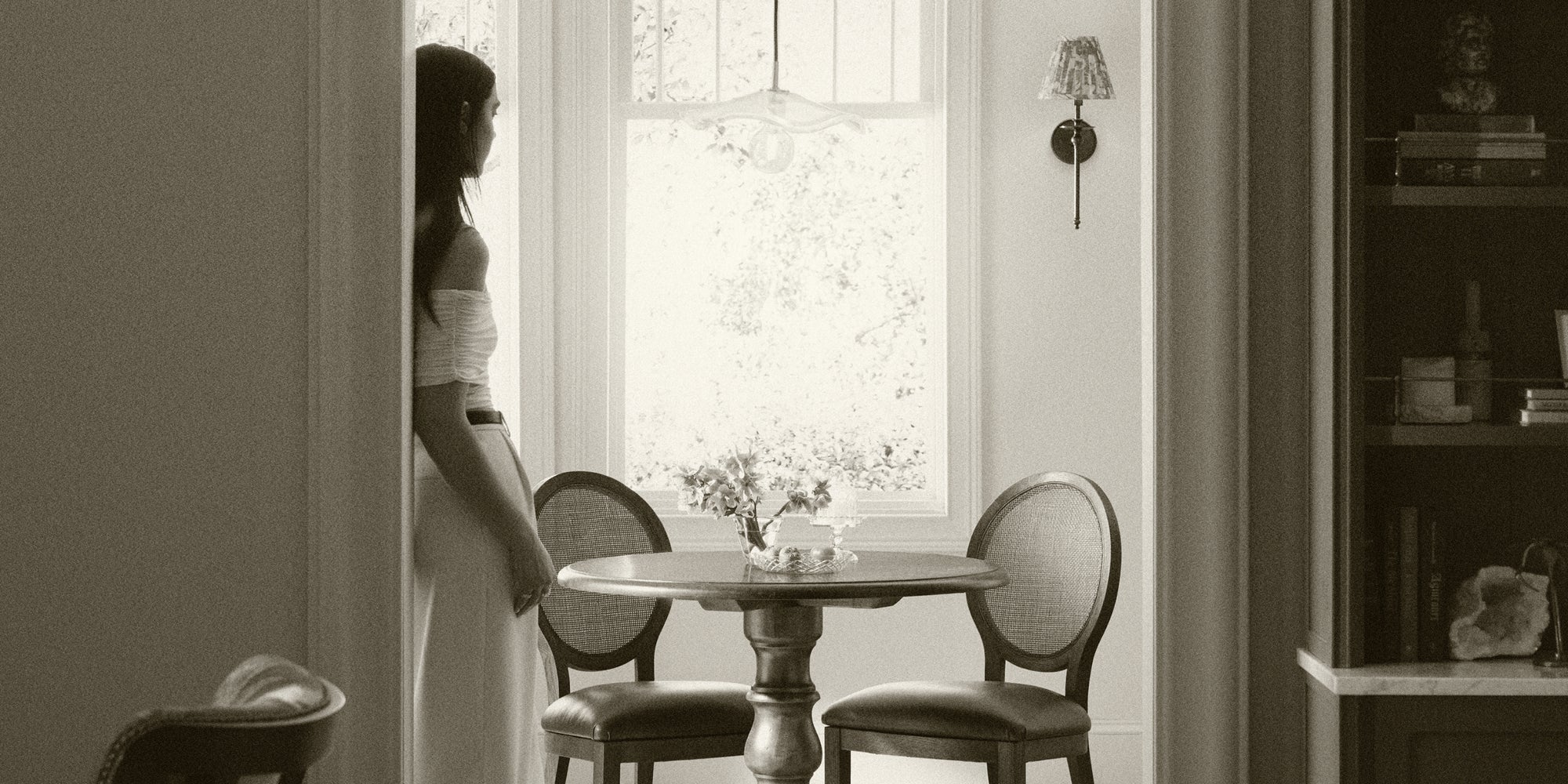 Person standing beside a traditional round dining table with upholstered dining chairs in a softly lit interior, styled with warm neutrals and timeless furnishings.
