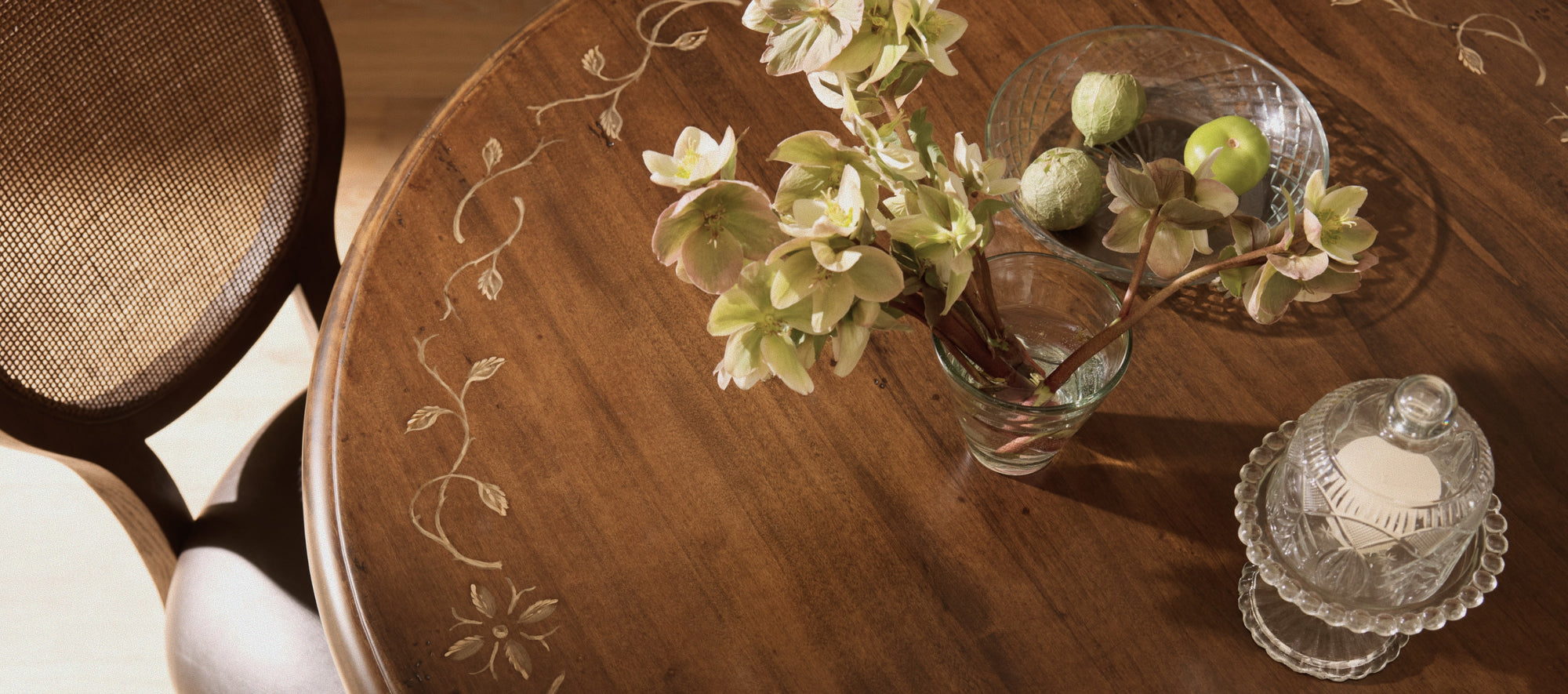 Round wood dining table with painted floral detailing, styled with fresh flowers, glassware, and decorative accents in warm natural light.