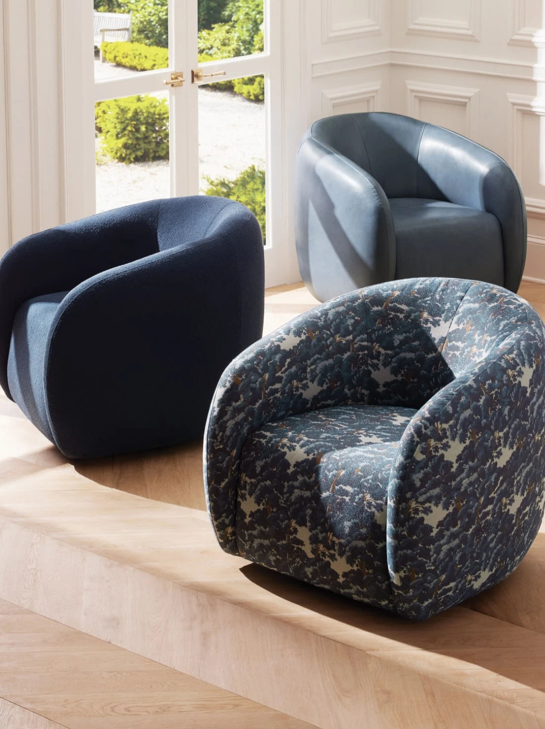 Three modern armchairs with a rounded, sculptural design are arranged in a bright room with tall white-trimmed windows. Each chair features a different upholstery: deep navy fabric, smooth slate blue leather, and a bold abstract forest-patterned textile, all resting on light wood flooring.