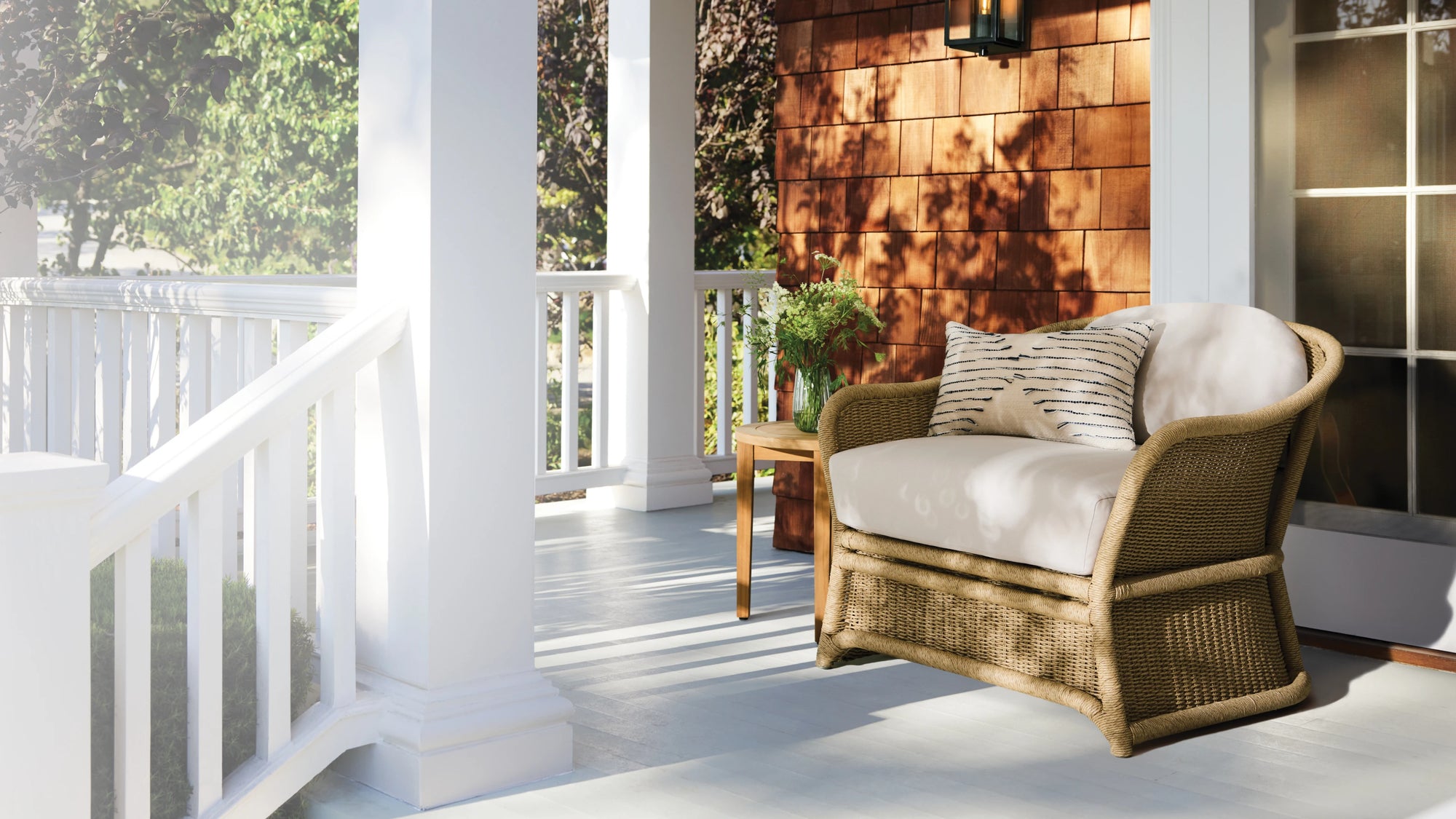 Cozy New England front porch with wicker chair, white cushions, striped pillow, and potted plant on a wooden side table.