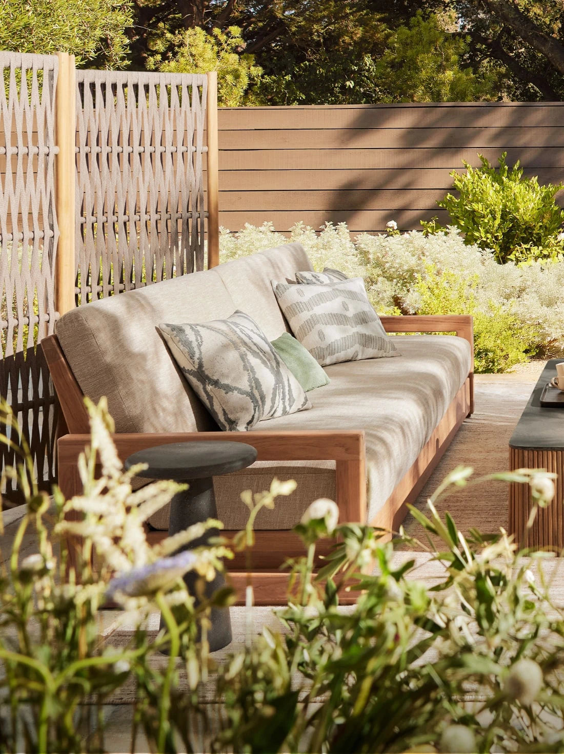 A serene patio scene showcasing sustainable outdoor furniture, including a wood-framed sofa with neutral cushions and earth-toned patterned pillows. Surrounded by soft greenery, flowering plants, and a woven privacy screen, the space blends eco-conscious design with natural beauty.