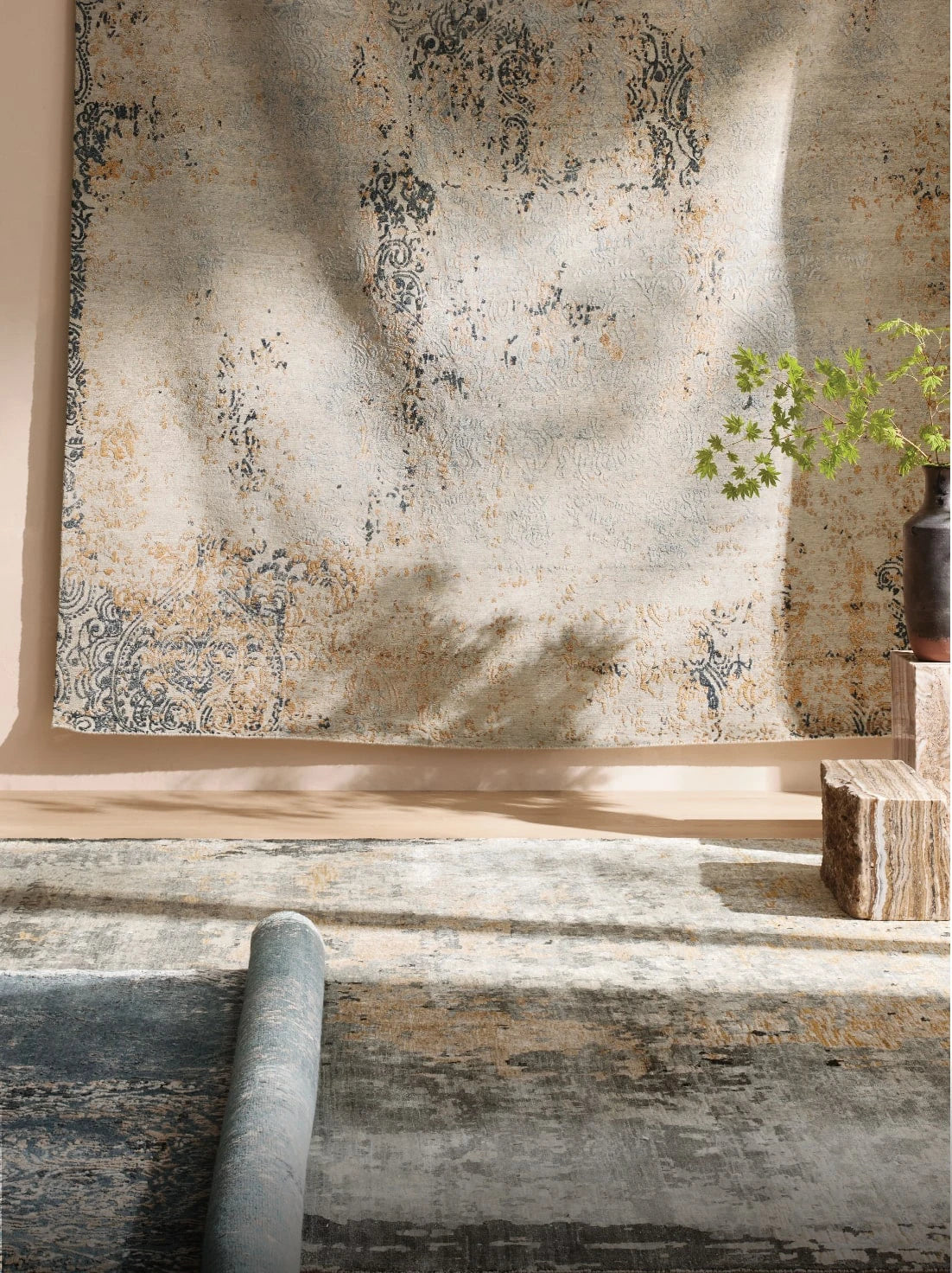 A pair of softly patterned area rugs in warm neutral tones with faded blue and golden ochre accents, styled in natural light. One rug is hung on the wall like a tapestry, while another is partially unrolled on the floor next to a stacked stone pedestal and a vase with leafy greenery.