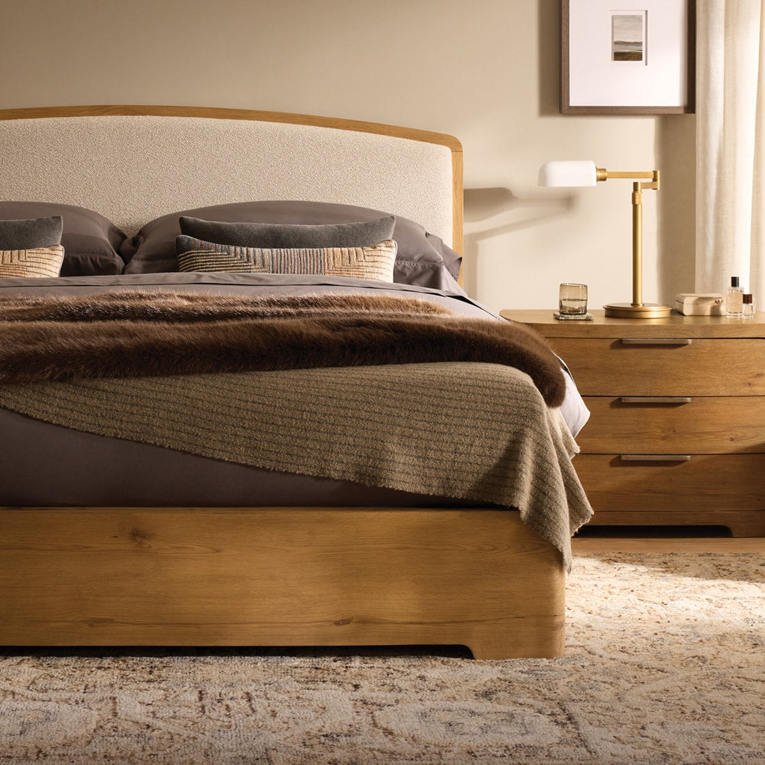 Bedroom with a curved upholstered and warm wood bed, layered neutral bedding, and warm wood nightstands, styled with soft lighting and rich seasonal textures.