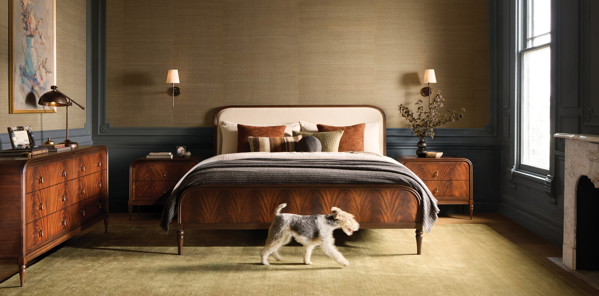 Bedroom with rich warm wood furnishings, layered bedding, and warm autumn accents, with a dog walking across the room in soft natural light.