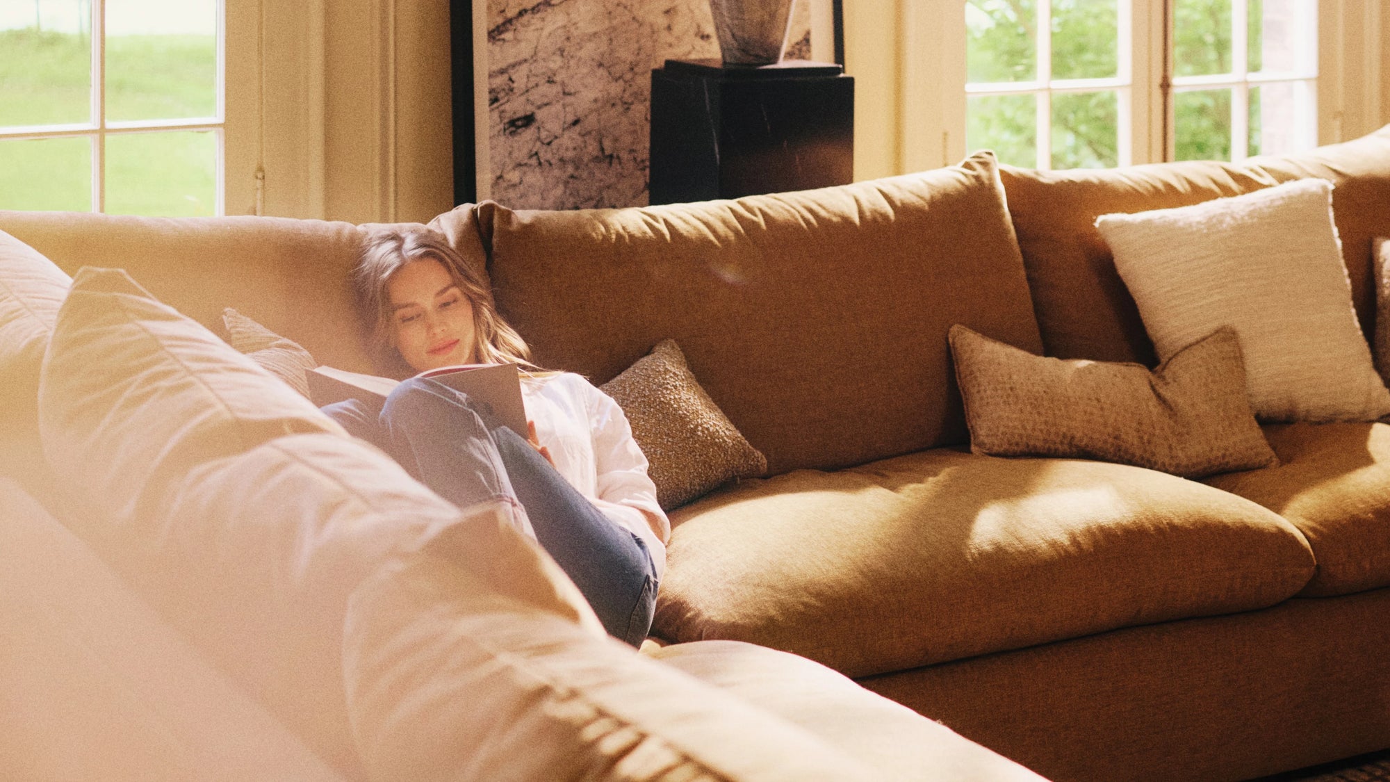 A woman sits comfortably on a large tan sectional sofa, reading a book with sunlight streaming through tall windows behind her. Soft beige and textured pillows surround her, creating a cozy and relaxed atmosphere.