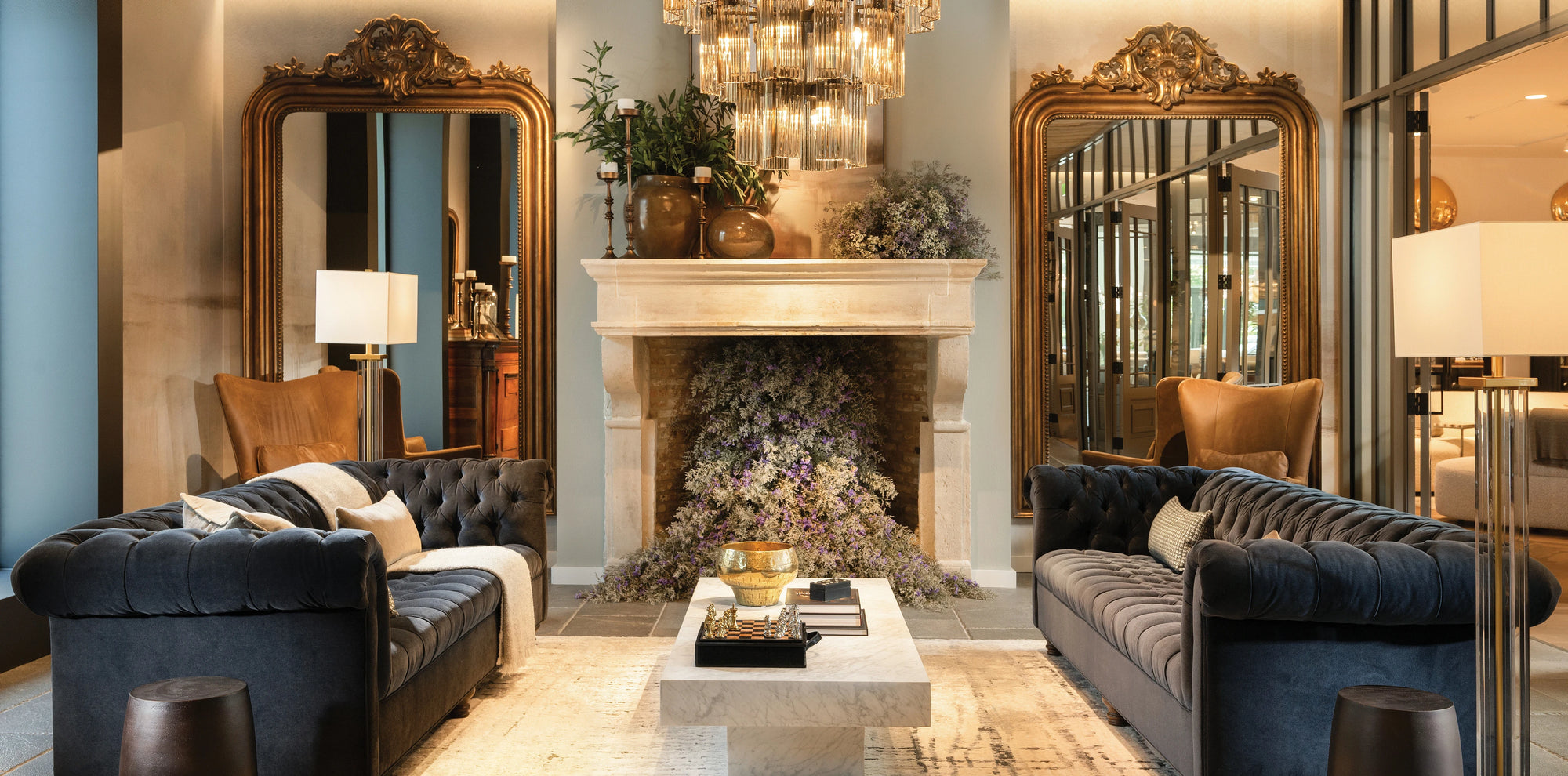 Luxurious Arhaus showroom featuring two navy tufted velvet sofas facing each other across a marble coffee table topped with decor books, a chess set, and a gold bowl. The space is styled with tall brass-framed mirrors, leather armchairs, modern floor lamps, and a grand crystal chandelier, all against a backdrop of neutral tones and lush greenery.
