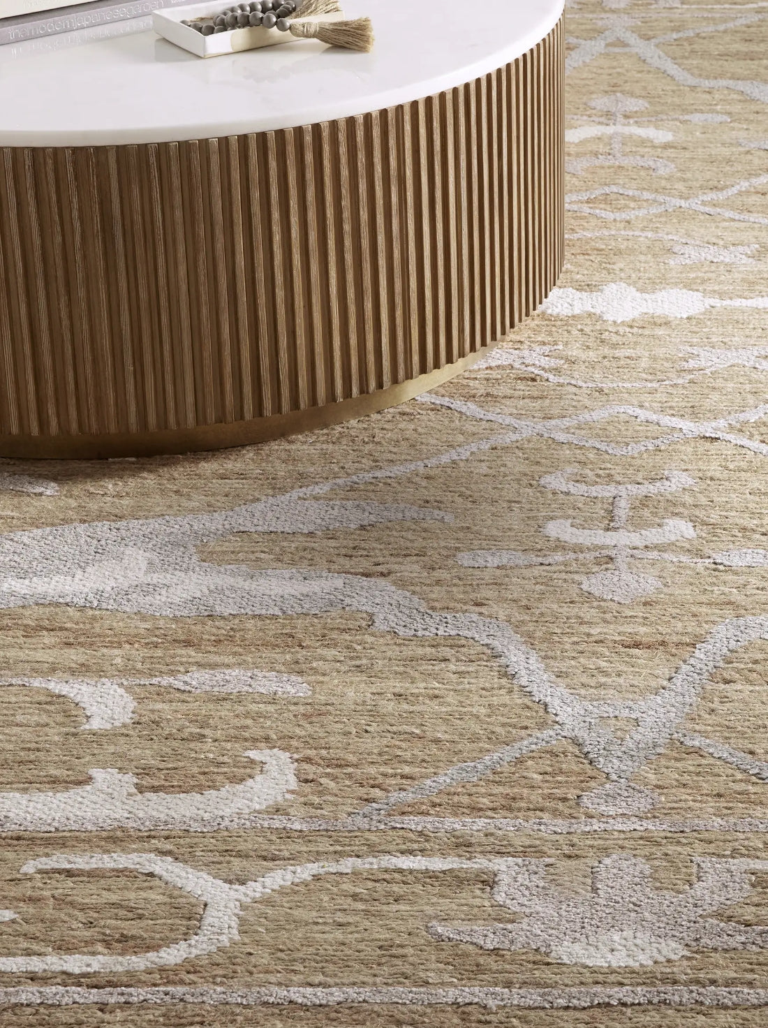 A round wooden coffee table with vertical grooves sits on a beige rug with elegant white patterns. Minimalist decor with light, earthy tones.