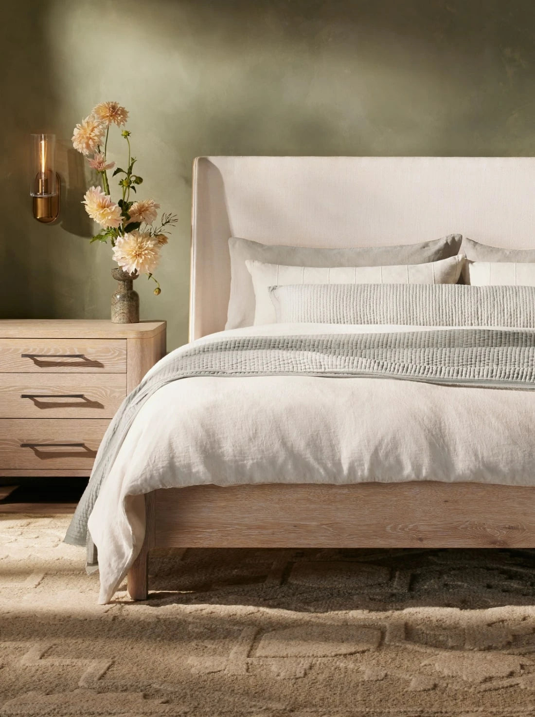 Bedroom with an upholstered bed, layered neutral bedding, and light wood nightstand, styled in soft tones with warm natural textures.