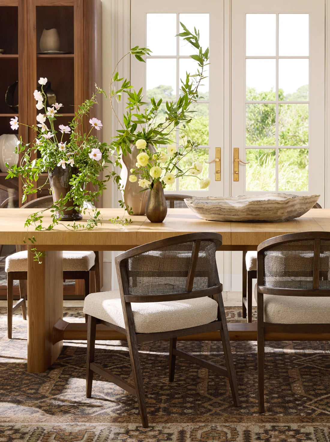 Dining room with a light wood table, woven-back chairs, and fresh floral arrangements, styled in a bright, sunlit interior.