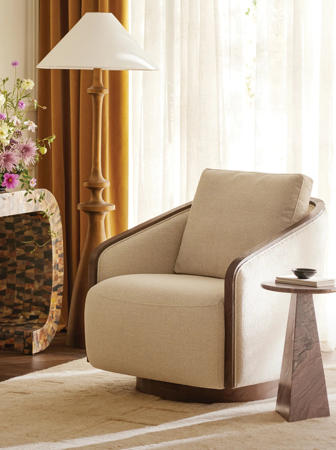 Living room with a curved upholstered chair, sculptural floor lamp, and warm wood accents, styled in a soft neutral palette with layered textures.